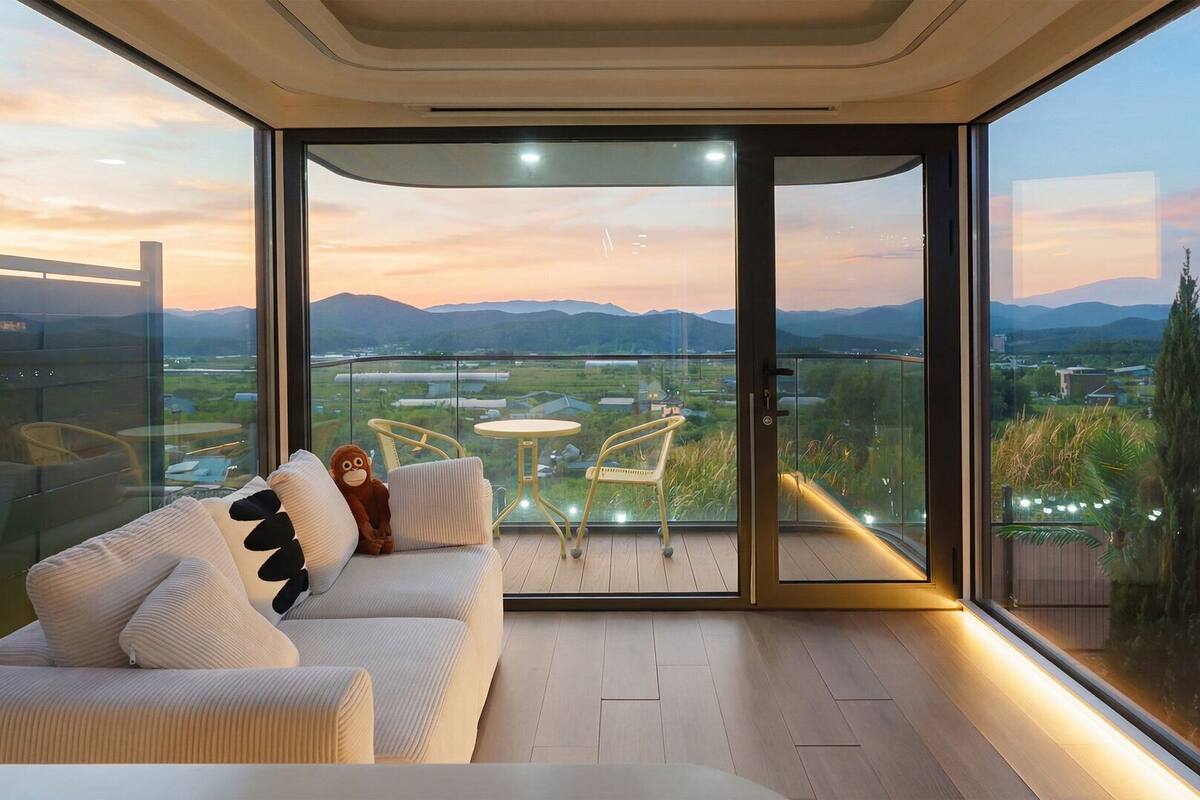 A modern living room with a sofa, two chairs, and a small table. A large sliding glass door leads to a balcony with a view of mountains and a cityscape.