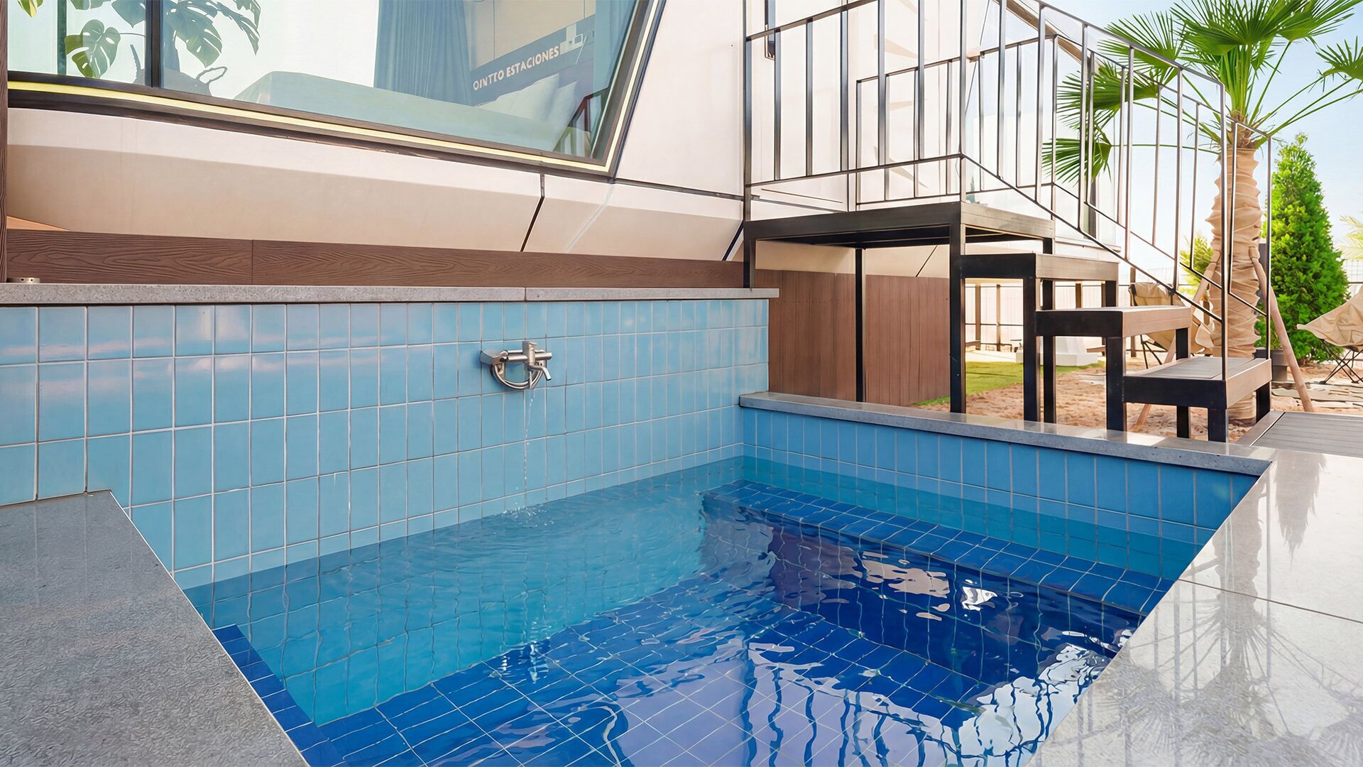 Una piscina con azulejos azules, una terraza de madera y vistas a un edificio moderno con amplios ventanales.
