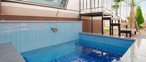 Una piscina con azulejos azules, una terraza de madera y vistas a un edificio moderno con amplios ventanales.