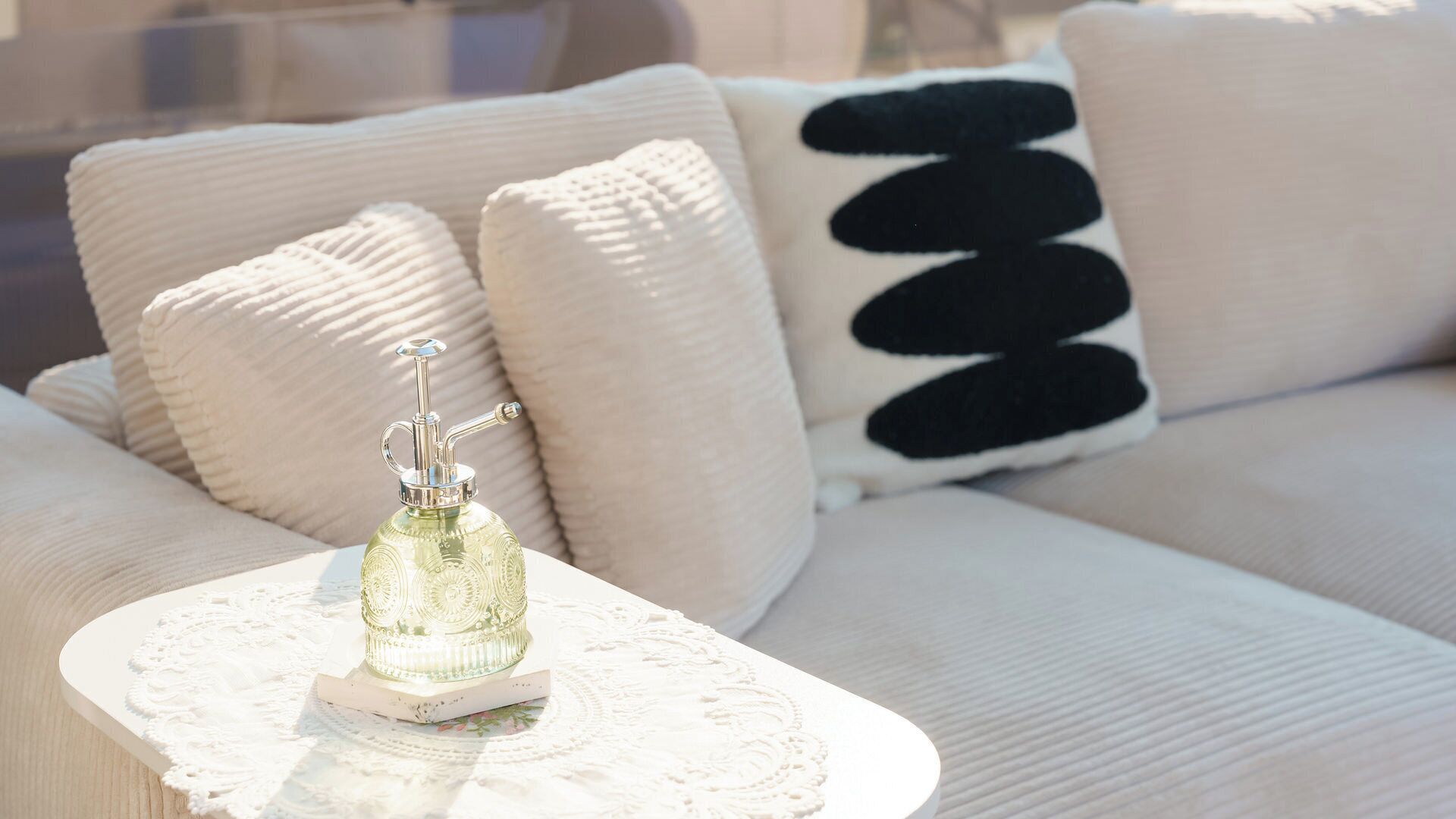 Un sofá blanco con un cojín texturizado, una mesa blanca con un mantelillo de encaje y una botella decorativa.
