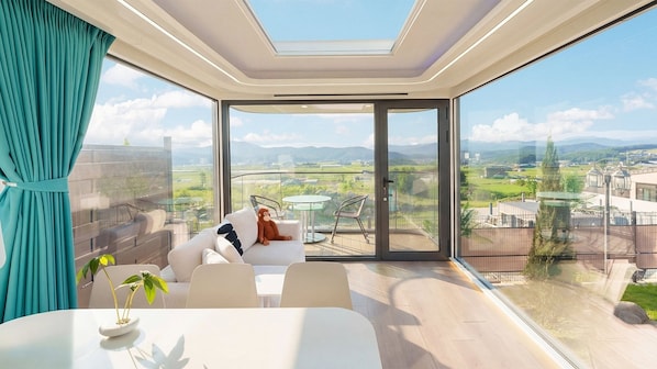 A modern room with a large sliding glass door leading to a balcony, a sofa, a dining table, and a view of a cityscape. - Gyeongju Holiday Space Pension (Gyeongju)