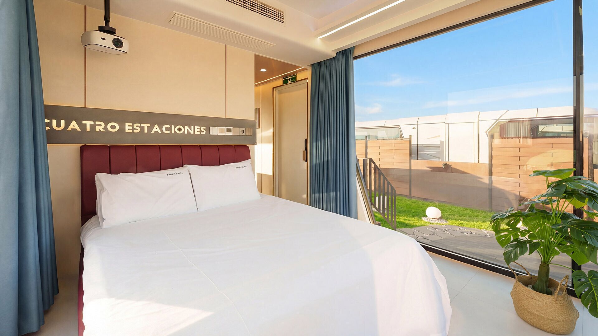 A hotel room with a bed, a projector, a view of the outside, and a sign with 'CUATRO ESTACIONES'.