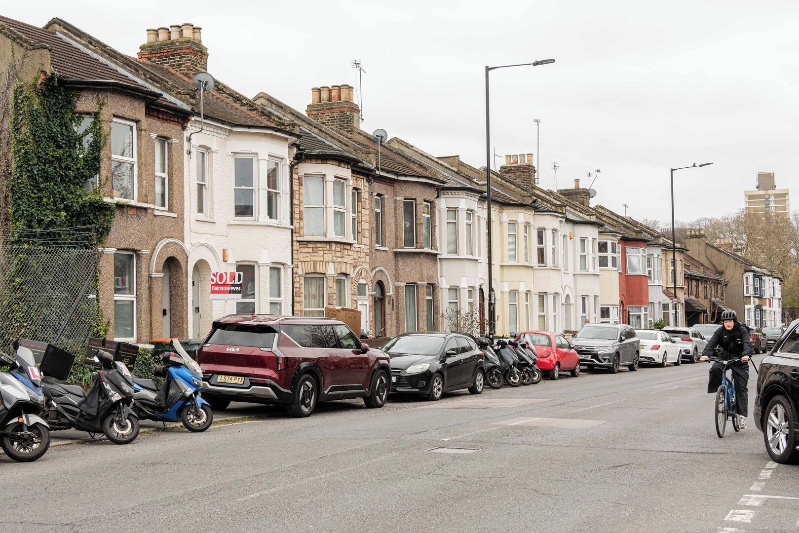A row of terraced houses with parked cars and motorcycles on the street.