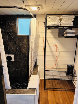 A bathroom with a shower, a towel rack with folded towels, a heater, and a power outlet.