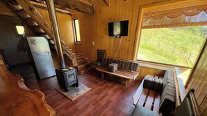 A wooden cabin interior with a wooden staircase, a small kitchen with a refrigerator, a fireplace, a sofa, and a television. - Secluded mountain cabin: steps to hot spring, river ,& lake (Futrono)