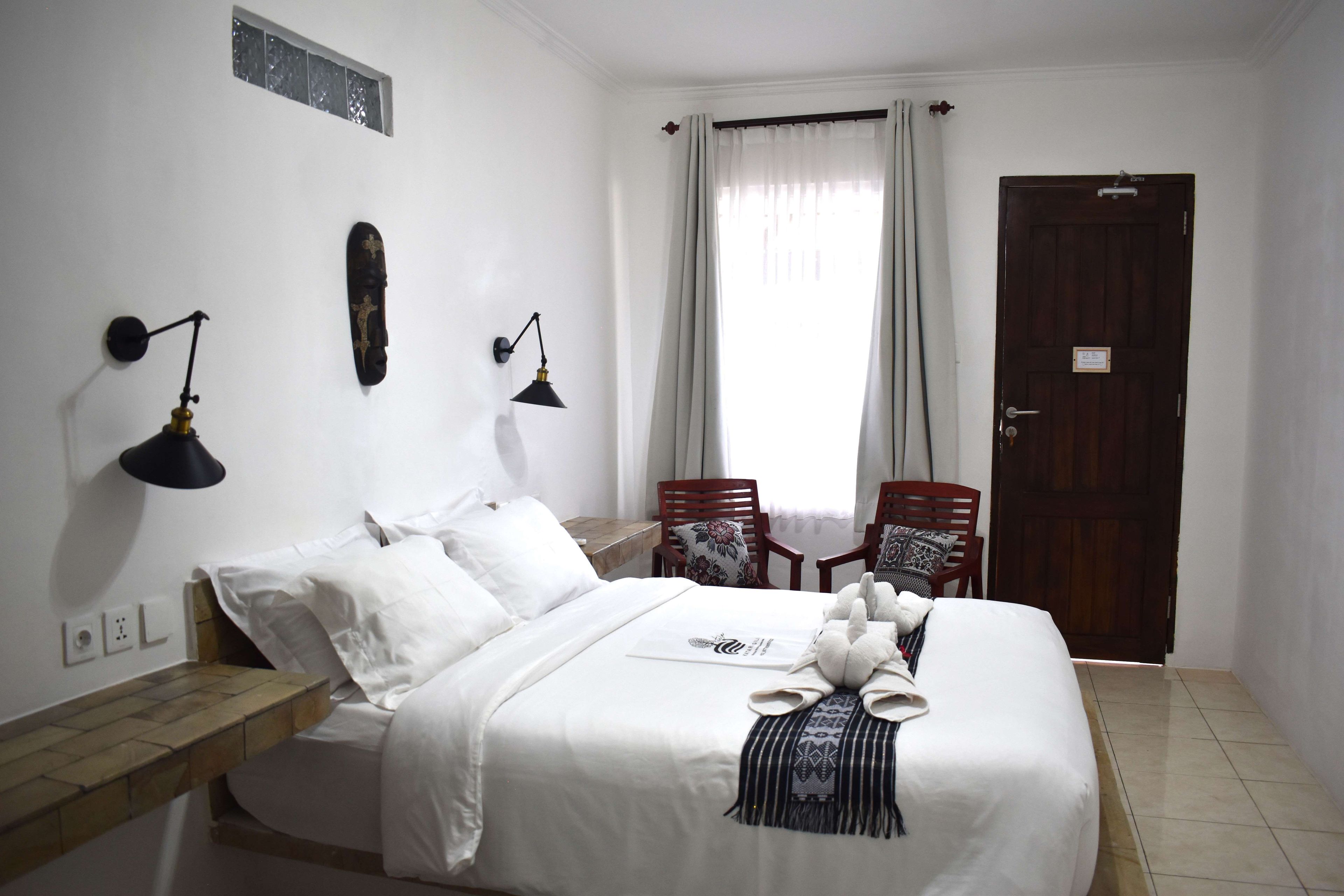 A hotel room with a bed, two bedside lamps, a wooden nightstand, a window with curtains, a door, and two chairs.