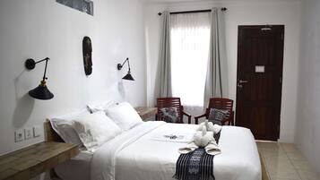 A hotel room with a bed, two bedside lamps, a wooden nightstand, a window with curtains, a door, and two chairs.