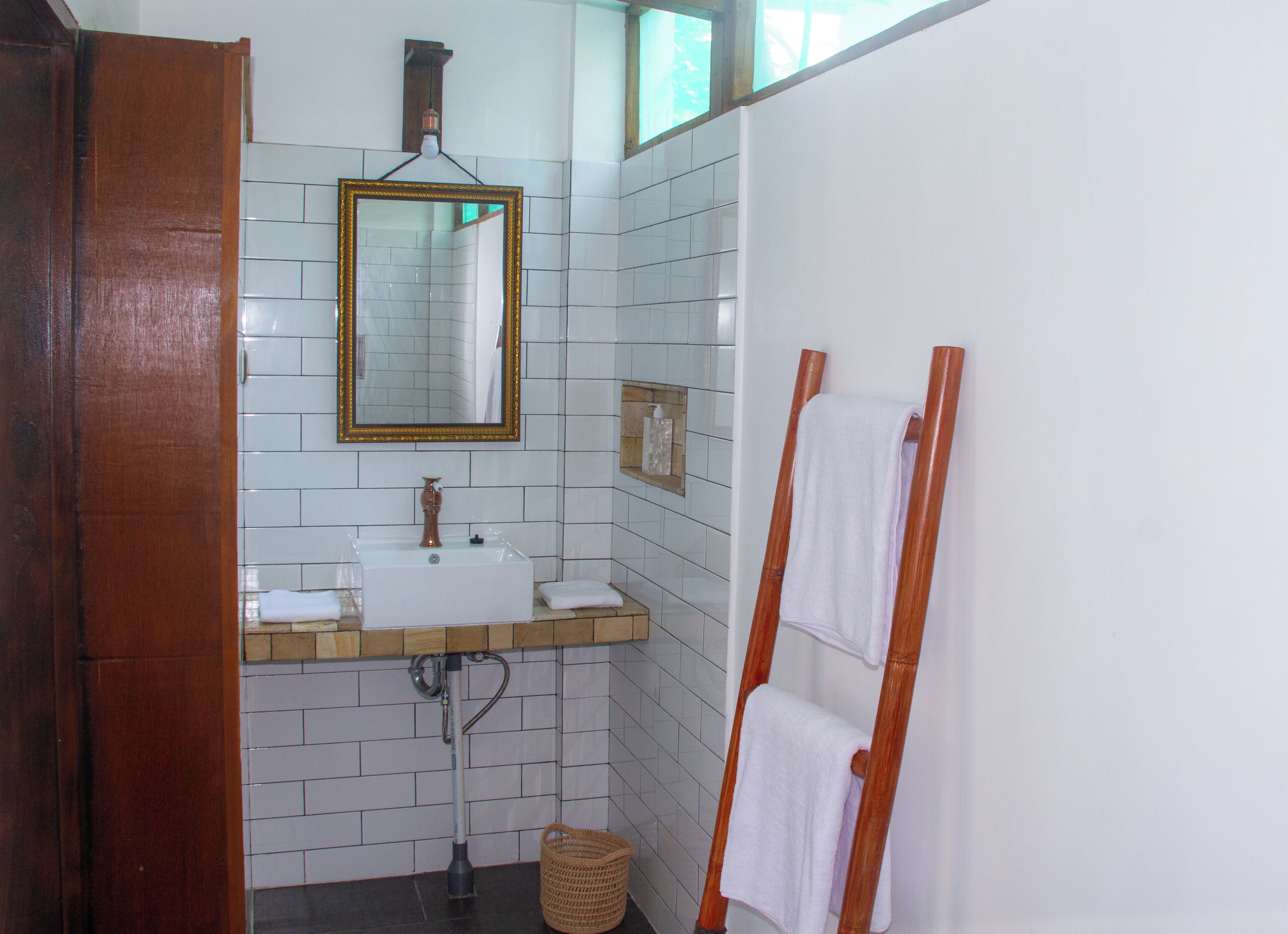 Um banheiro com parede revestida de azulejos brancos, toucador de madeira com pia, espelho com moldura dourada e porta-toalhas de madeira.