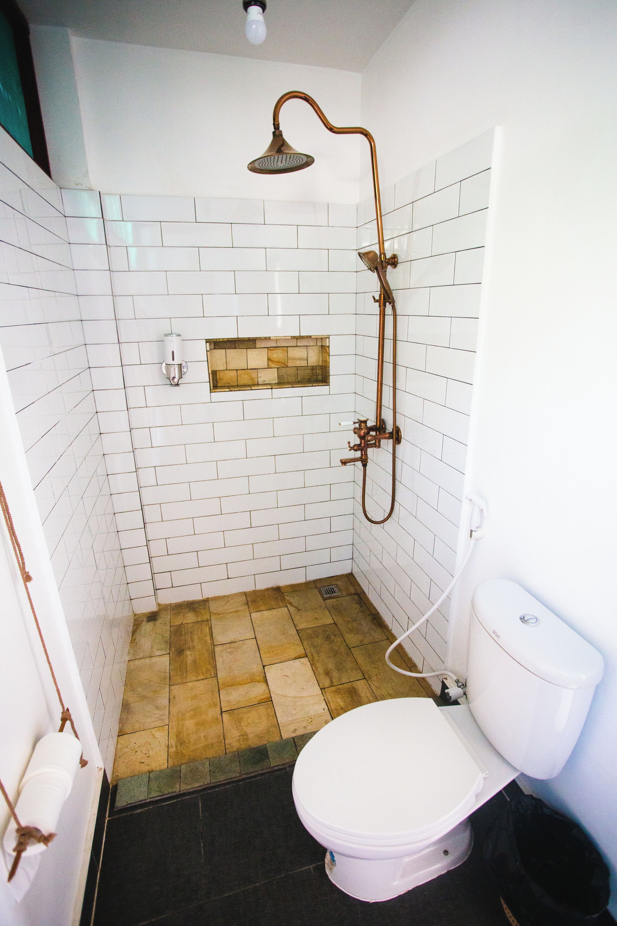 Une salle de bain avec des murs carrelés de carreaux blancs, un plancher en bois, des toilettes blanches et une douche dotée d’un pommeau de douche incurvé.