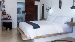 A bedroom with a bed, a wooden headboard, a bedside table, a lamp, a mirror, and a bathroom visible through an open door. - Katara Gili Boutique Rooms and Bungalows (Gili Trawangan)