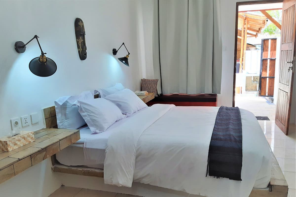 Ein Schlafzimmer mit Bett, Nachttischen, einer Wandlampe und Blick ins Freie.