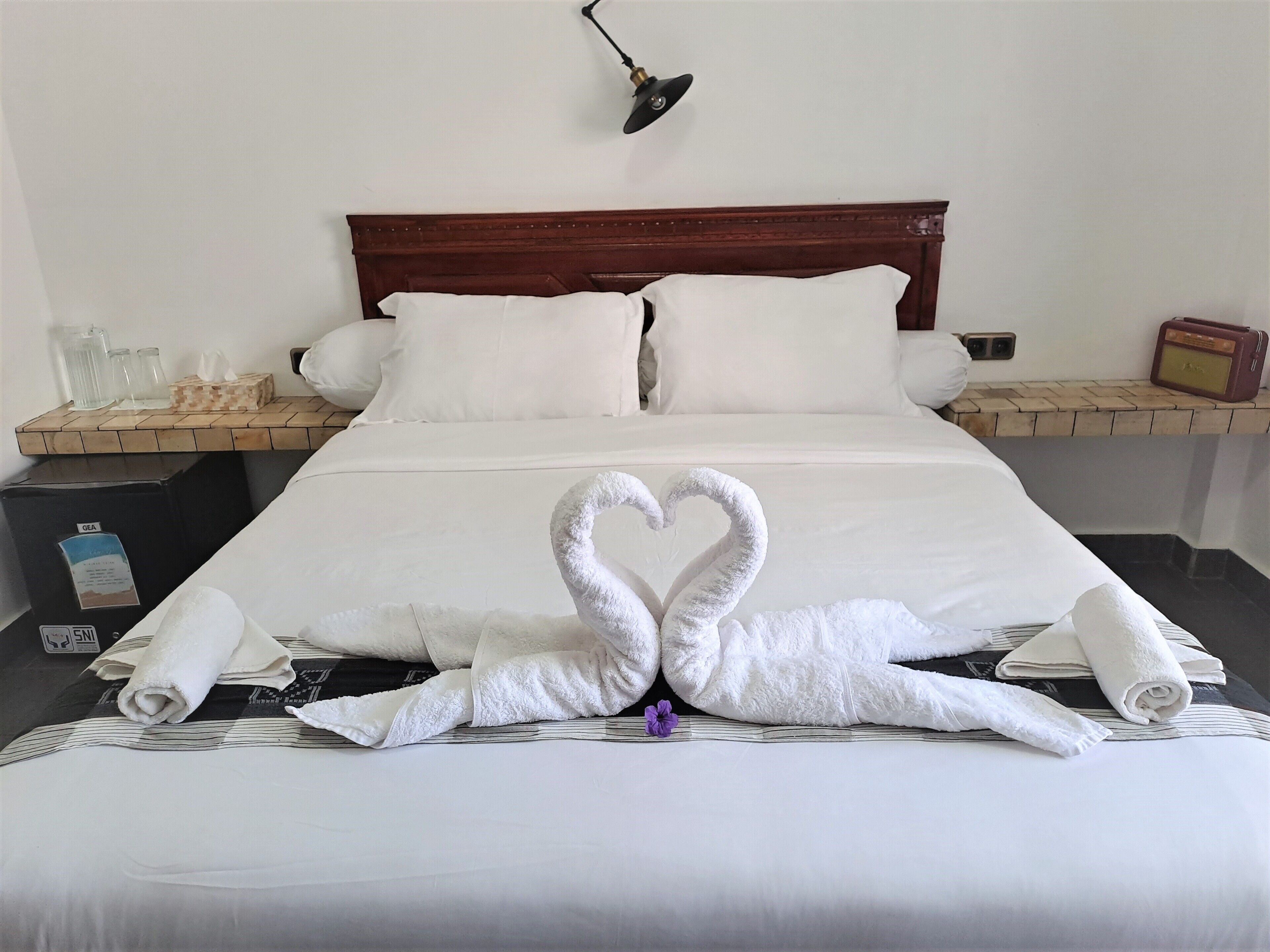 A hotel room with a neatly made bed, a bedside table with a radio, and a wall-mounted lamp.