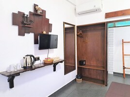 Una habitación con una televisión montada en la pared, un estante de madera con una tetera, tazas y una cesta, y un armario de madera con espejo.