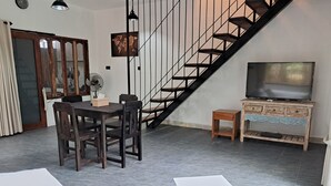 A living room with a dining table, chairs, a white coffee table, a TV on a wooden stand, a staircase, and a world map on the wall. - Katara Gili Boutique Rooms and Bungalows (Gili Trawangan)