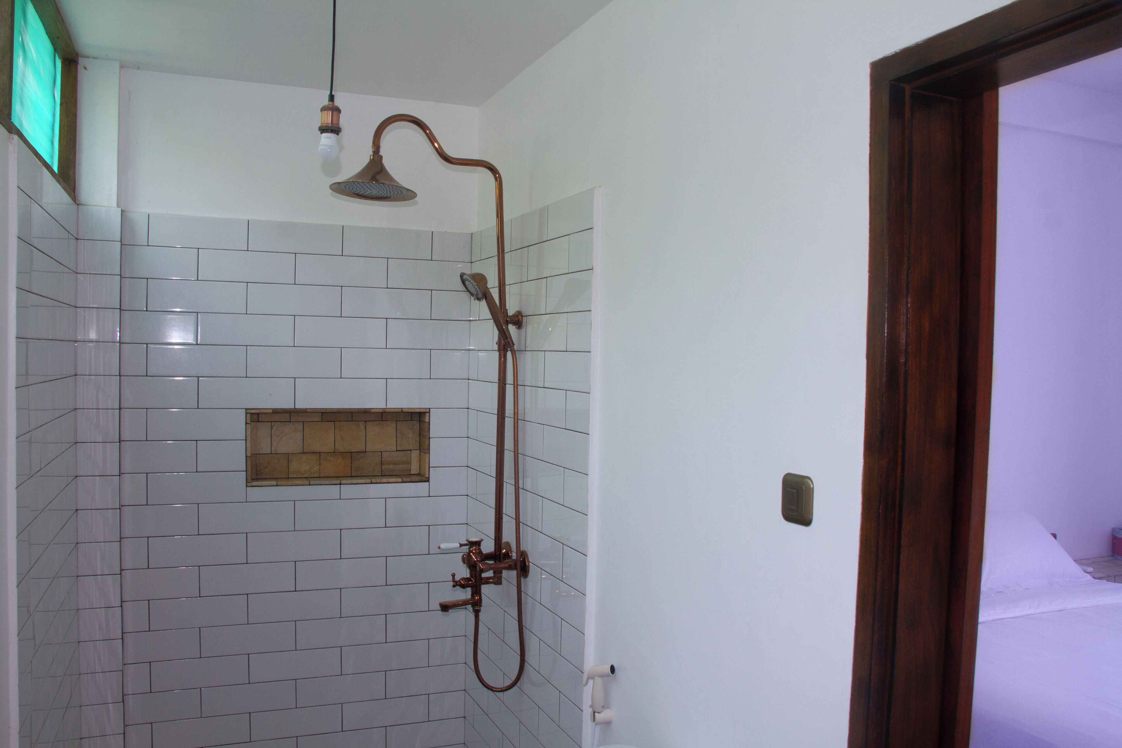 Casa de banho com paredes revestidas a mosaico branco, chuveiro com um esguicho curvo e porta de madeira.