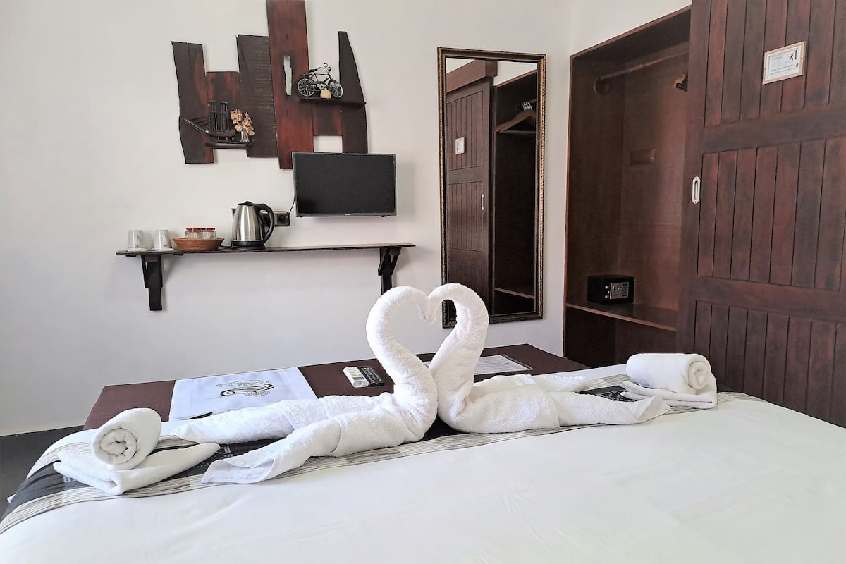 Ein ordentlich bezogenes Bett mit einer Schwan-Towel-Motivs, ein an der Wand befestigter Fernseher, eine Holztür und ein Regal mit Wasserkocher und Tassen.