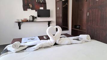 A neatly made bed with a swan towel design, a television on the wall, a wooden door, and a shelf with a kettle and cups.