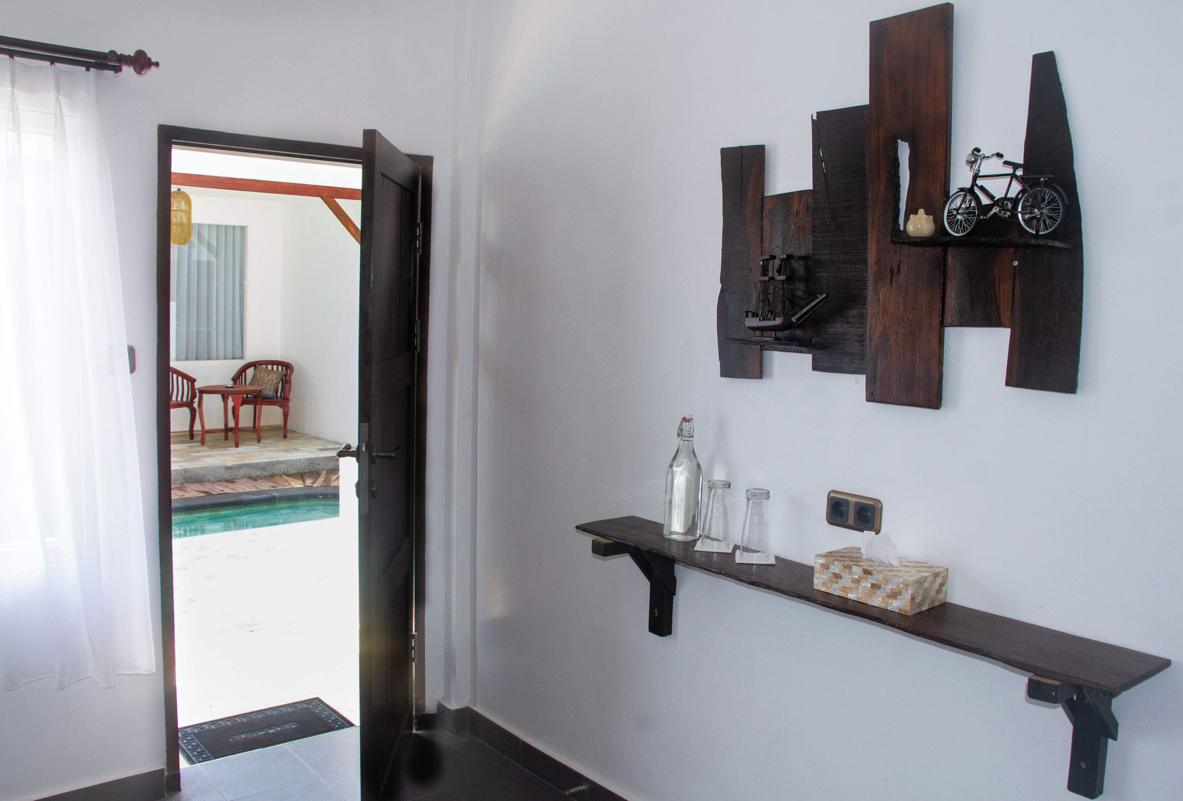 Una habitación con una puerta que da a un balcón, un estante de madera con botellas y pañuelos, y un estante en la pared con una bicicleta y un adorno.
