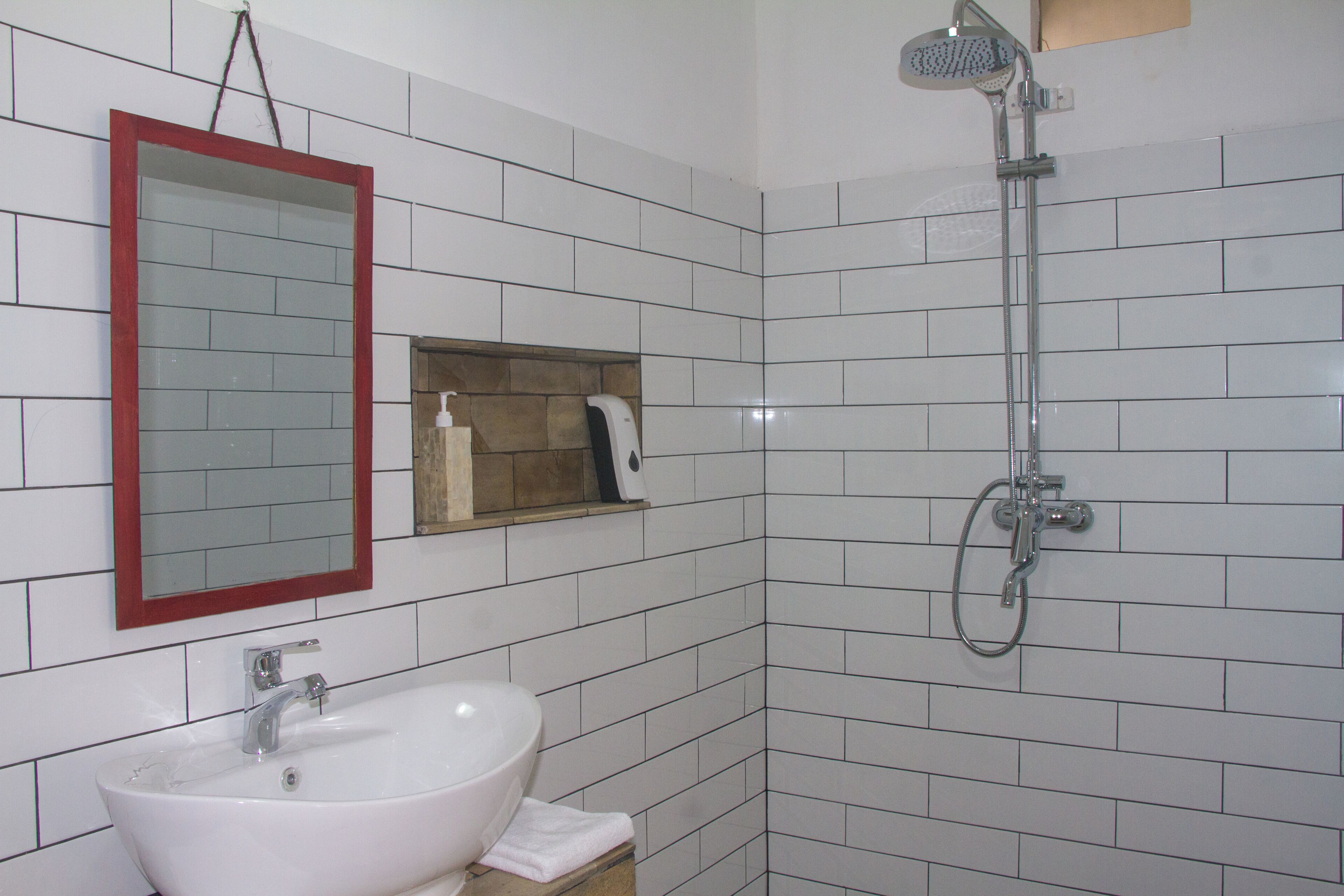 A bathroom with a white sink, a mirror with a red frame, a handheld showerhead, and a soap dispenser.