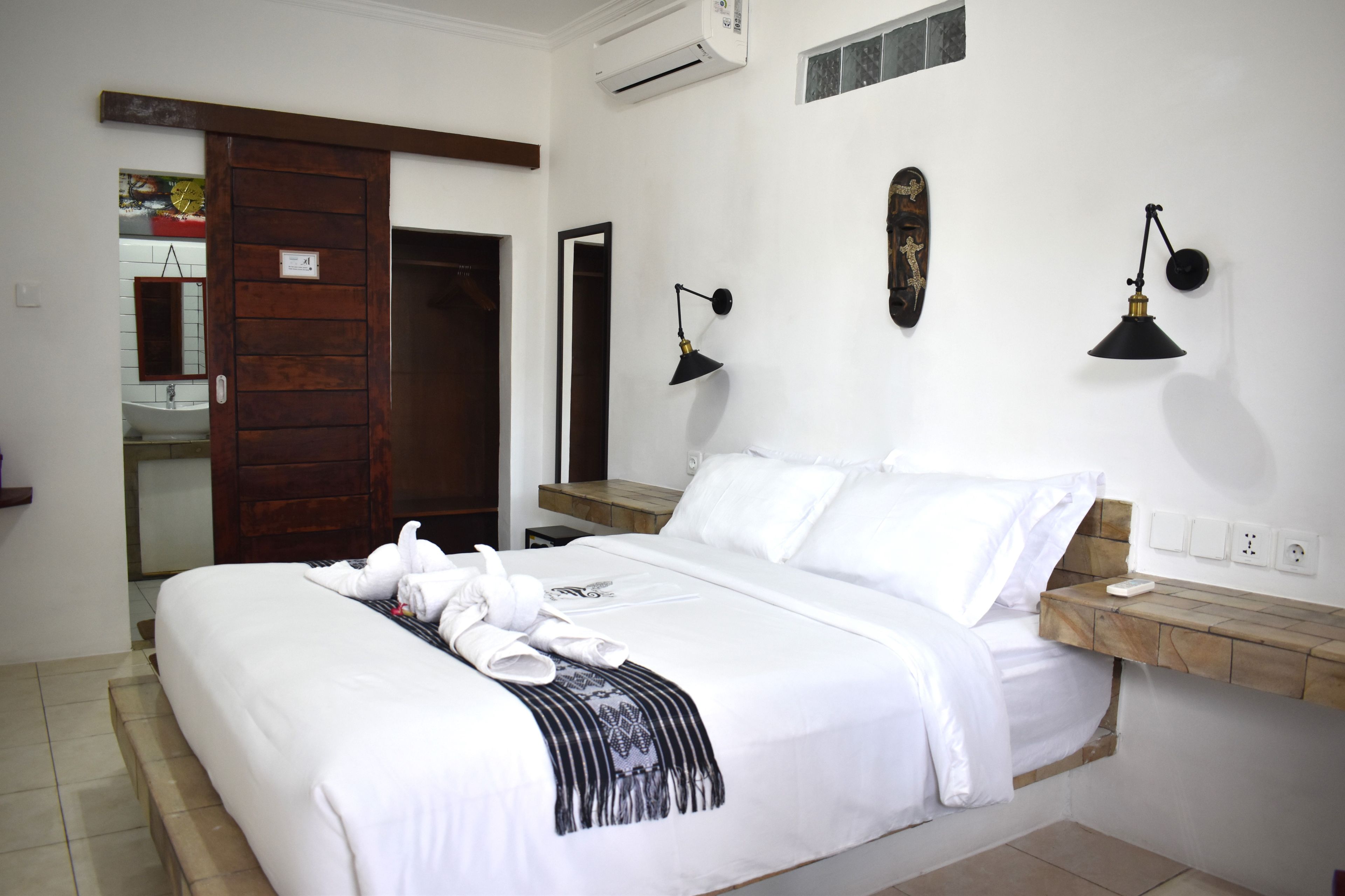 A hotel room with a bed, a wooden door, a mirror, a wall-mounted air conditioner, and a bedside table with a lamp.