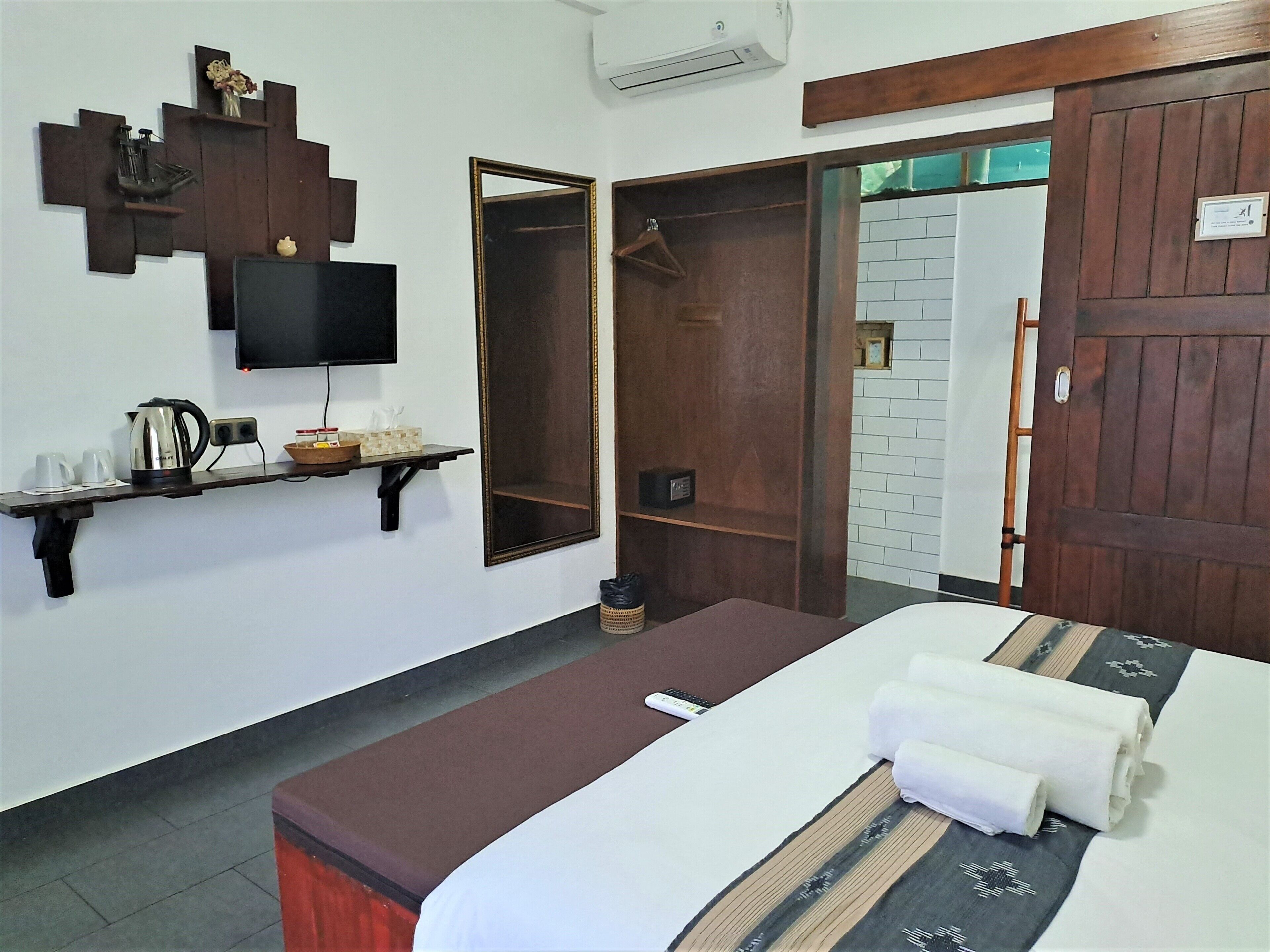 Habitación de hotel con cama, televisor montado en la pared, estante de madera con objetos y puerta que da a otra habitación.
