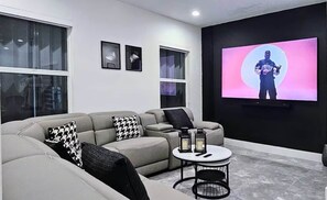 Ein modernes Wohnzimmer mit einem grauen Sofa, Kissen in Schwarzweißmuster, einem kleinen runden Tisch mit zwei Kerzen und einem großen Fernseher, der eine Person zeigt.