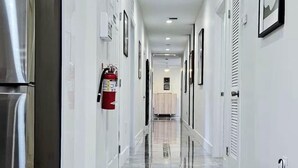 A corridor with a stainless steel refrigerator, a fire extinguisher, and framed pictures on the walls.