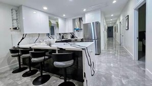 A modern kitchen with white cabinets, a central island with bar stools, stainless steel appliances, and a marble floor.