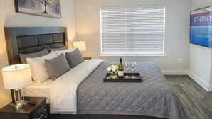 A bedroom with a bed, bedside lamps, a ceiling fan, a window with blinds, and a wall-mounted TV displaying a phone number. - Walk to Beach | Family-Friendly 3BR Home w/ Garage (Biloxi)