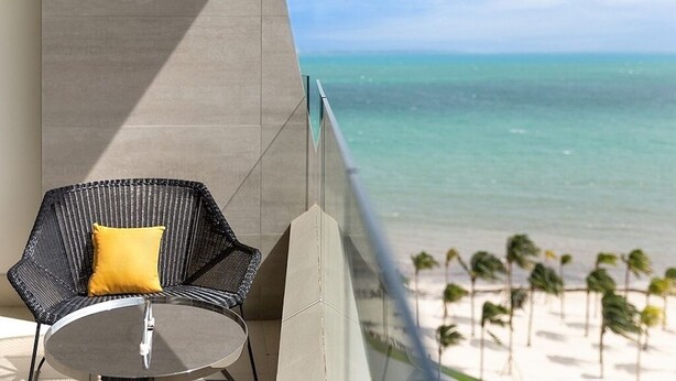 Ein Balkon mit Stuhl und Tisch, mit Blick auf einen Strand mit Palmen.