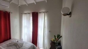 Ett hotellrum med en säng, röda gardiner, vita gardiner, en väggmonterad lampa och en liten vas med blommor.