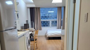 Una camera d'albergo compatta con angolo cottura, lavatrice, un letto e vista sulla città dalla finestra.
