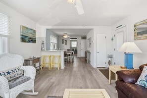 Ein Wohnzimmer mit einem weißen Korbstuhl, einem braunen Ledersofa, einer Küche im Hintergrund und einem Essbereich mit gelben Hocker.