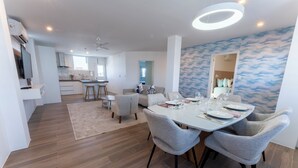 Una cocina moderna con armarios blancos, una zona de comedor con una mesa redonda y sillas grises, y un salón con sofá y televisor.