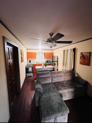 A living room with a sofa, a ceiling fan, and a kitchen in the background. - Located in Flowers Bay, enjoy what this 1-bedroom villa. (Flowers Bay)