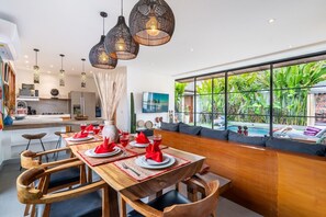 Ein moderner Essbereich mit Holztisch, gedeckt für vier Personen, einer Küche im Hintergrund und einem großen Fenster mit Blick auf einen Pool und Grünflächen.