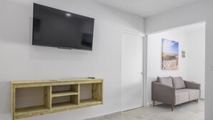 A modern living room with a flat-screen TV, a wooden console table, a grey sofa, and a beach-themed painting. - Charming 1BR1BA Getaway/Pool, BBQ &Laundry Access (Oranjestad)