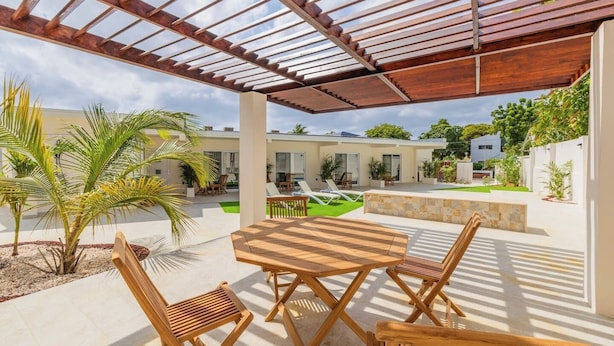A covered outdoor patio with a wooden table and chairs, a pool, and a view of a modern house in the background. - Charming 1BR1BA Getaway/Pool, BBQ &Laundry Access (Oranjestad)
