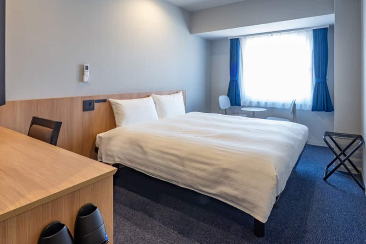 A hotel room with a bed, a desk, a chair, a small table, and a window with curtains.