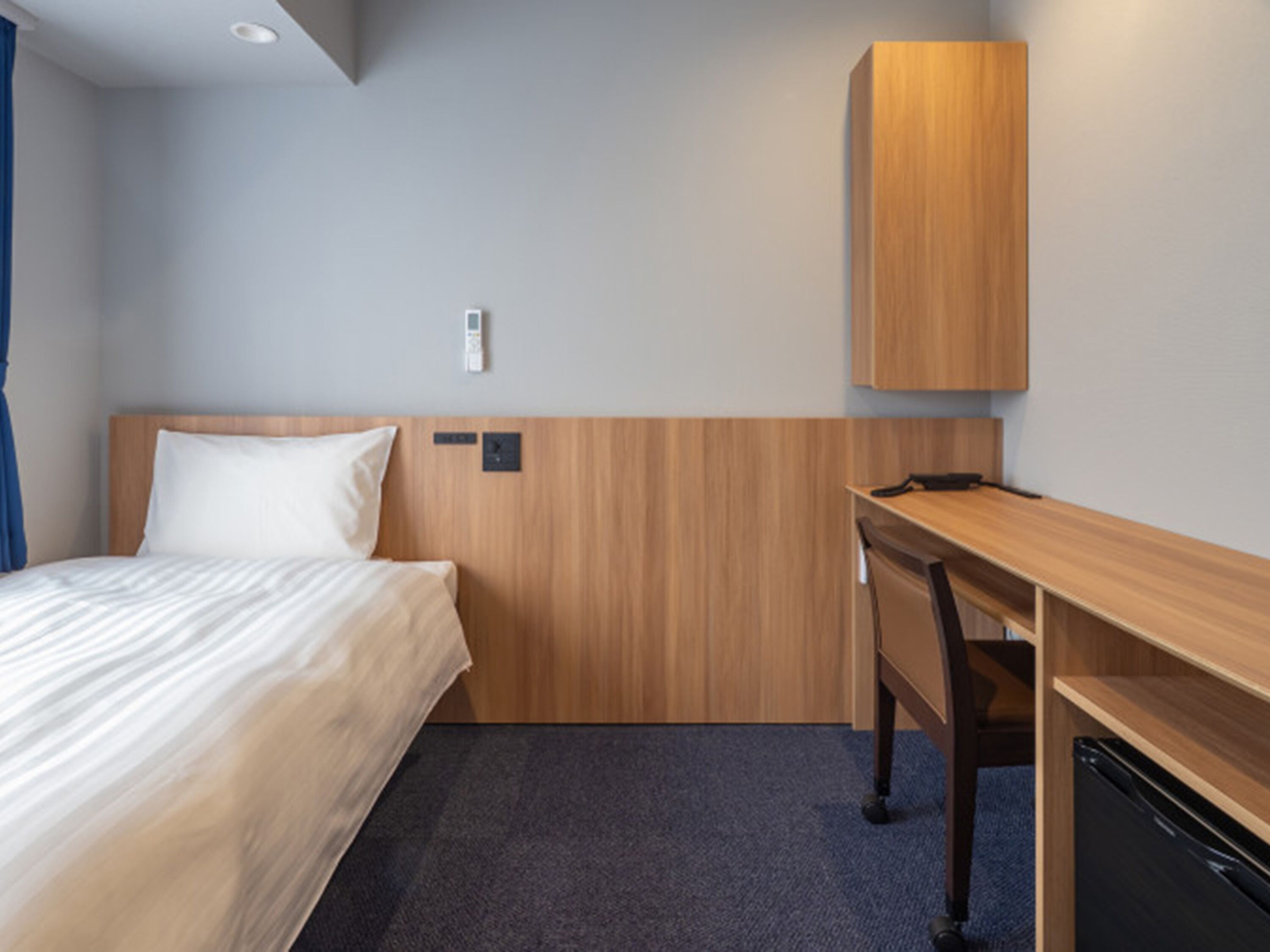 A hotel room with a bed, a desk, a wardrobe, and a wall-mounted control panel.