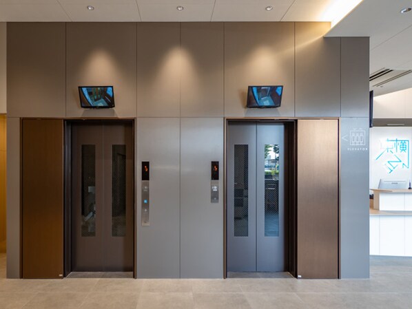 Dos puertas de ascensor con pantallas digitales, un mostrador de recepción y un cartel con flechas direccionales.