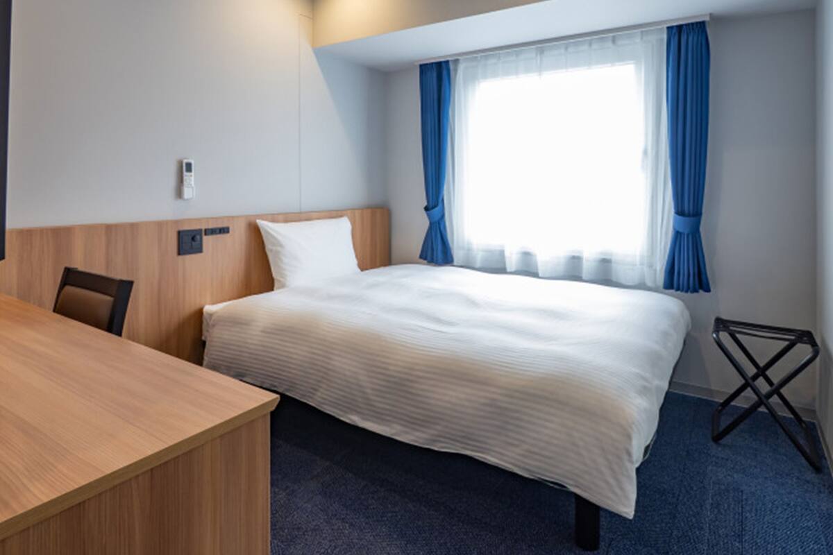 A hotel room with a bed, a wooden desk, a chair, a small table, and a window with curtains.