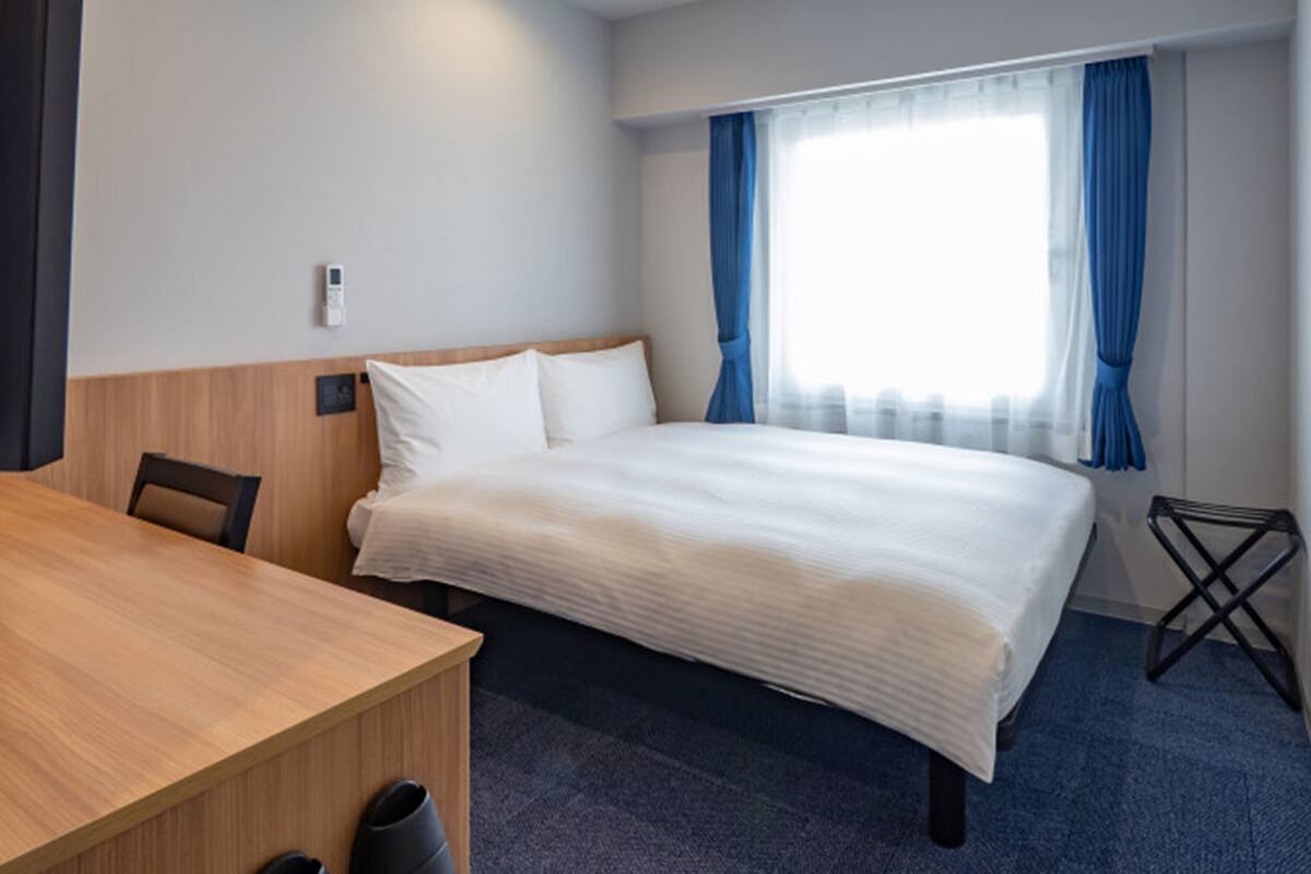 A hotel room with a bed, a wooden desk, a chair, and a window with curtains.