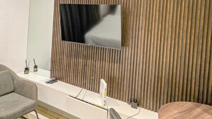 Una habitación de hotel moderna con un televisor de pantalla plana montado en una pared de paneles de madera, una mesita consola blanca, un sillón gris, una mesa redonda de madera y un equipo de aire acondicionado.
