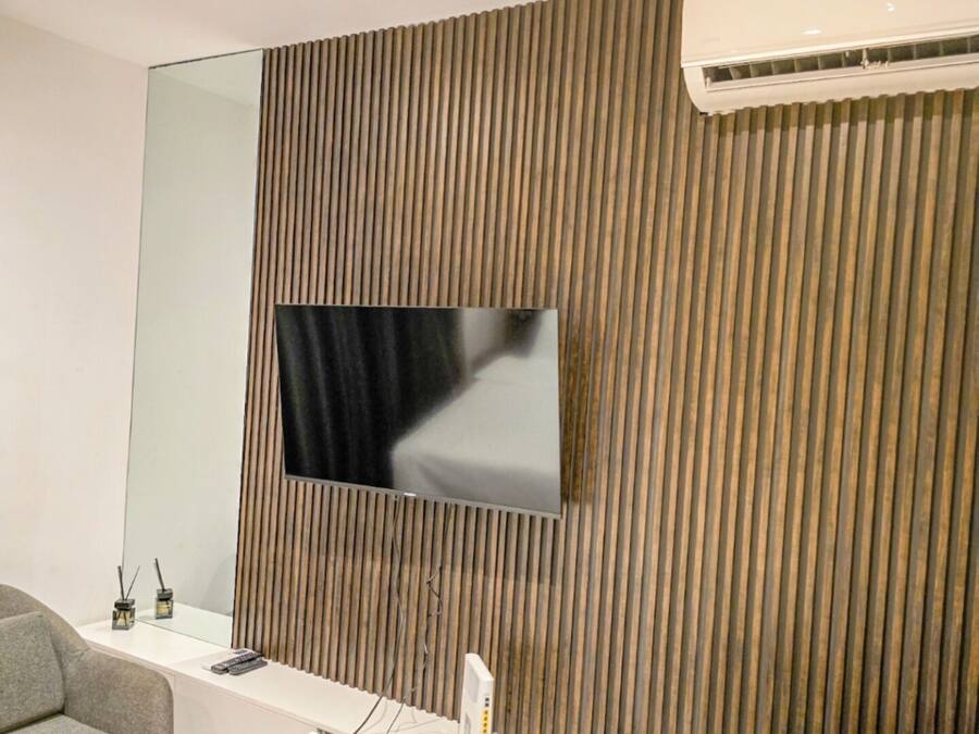 Ein modernes Hotelzimmer mit Flachbildfernseher an einer Wand aus Holzpaneelen, einem weißen Konsolentisch, einem grauen Sessel, einem runden Holztisch und einer Klimaanlage.