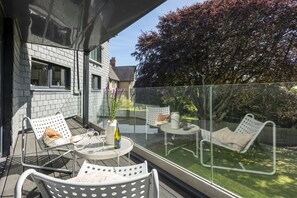 A balcony with outdoor seating, a glass barrier, and a view of a tree and house.