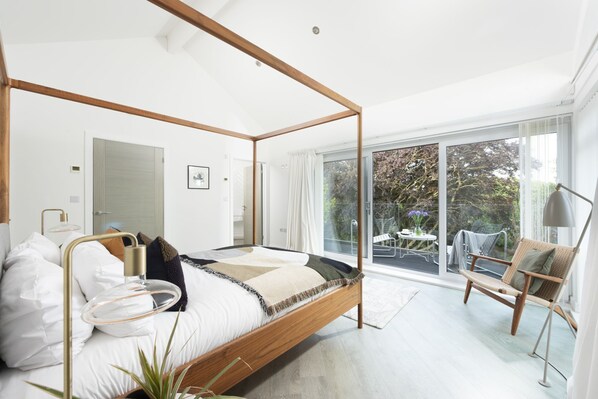 Un dormitorio moderno con cama con dosel, una zona de estar con una silla y una mesa, y un balcón con vistas a árboles.