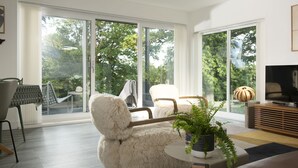 A modern living room with a large sliding glass door, a white furry chair, a round coffee table with a plant, and a TV on a wooden stand. - Estuary Watch - River view, Pets welcome, Family Friendly (Lelant)