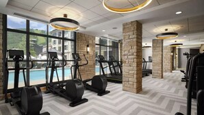 A gym with treadmills, elliptical machines, and stationary bikes, large windows with a view of a pool and buildings, and stone pillars. - Ski-in/Ski-out Park Hyatt Beaver Creek | Luxury Double Villa (Avon)