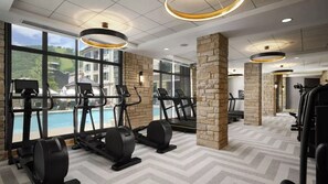 Une salle de sport équipée de tapis de course, d’appareils elliptiques et de vélos d’appartement, de grandes fenêtres offrant une vue sur une piscine et des bâtiments, ainsi que de piliers en pierre.